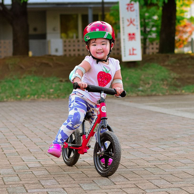 A child in a purple bike helmet rides a green Strider 12 Sport with a bridge and mountains in the background