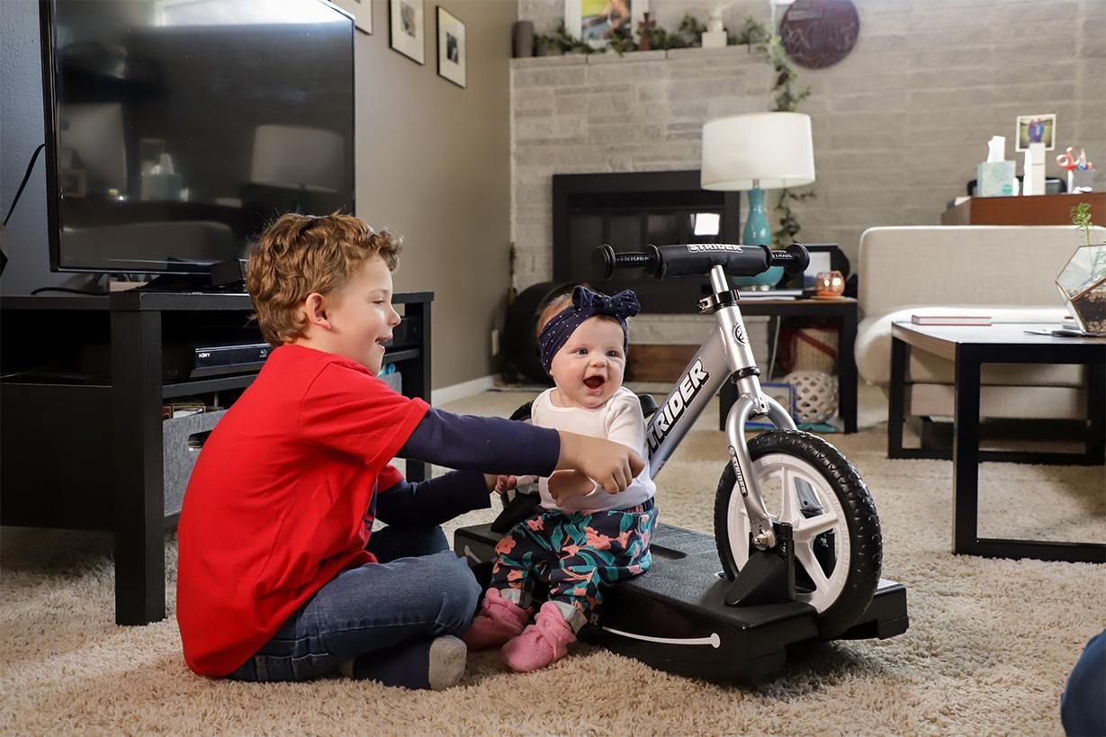 An older brother holds up his little sister on a silver Strider Rocking Bike